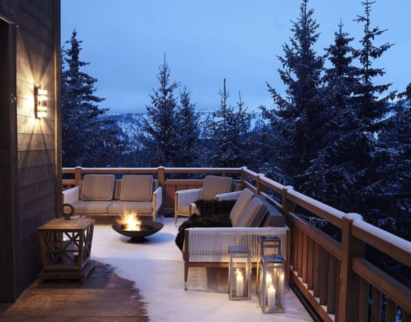 A chalet terrace in the snow at dusk, with lanterns and a fire bowl among the firs