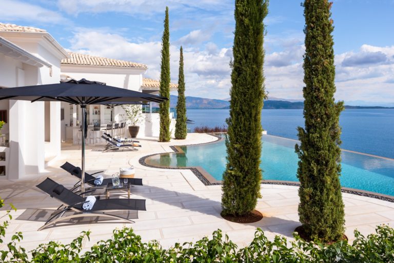 The pool of Villa Ultima Corfu between tall cypresses, with the Ionian Sea beyond
