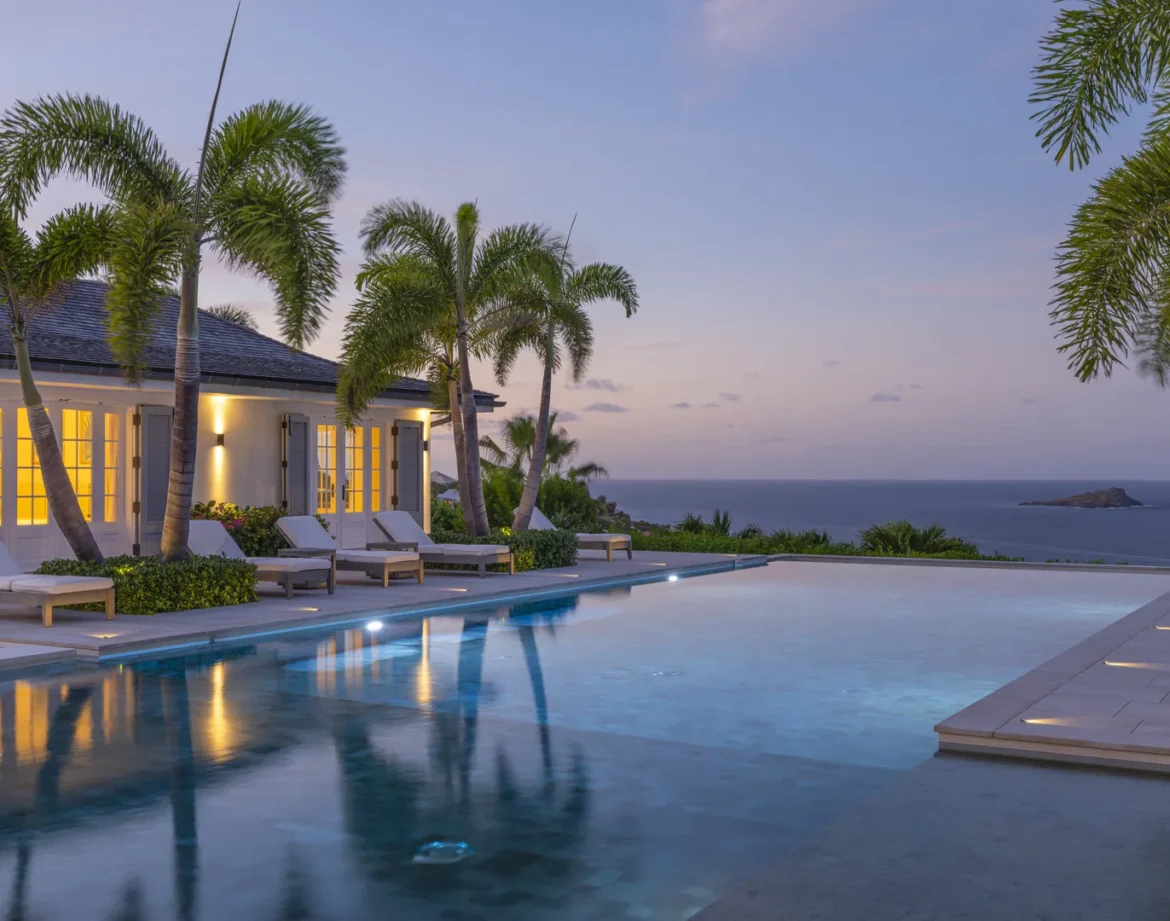 St Barts Villa Rentals – Luxury Villas in St Barths | Haute Retreats 10 a pool with palm trees and a house with a beach in the background in st barts - st barts villa rentals