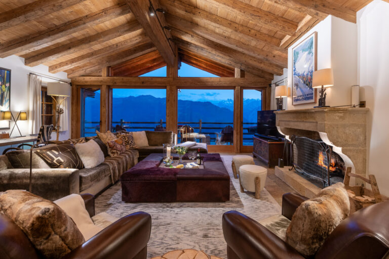 A chalet living room under a timber roof, with the fire lit and the mountains through the windows